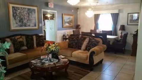 A cozy living room area with two beige and brown sofas arranged around a dark wooden coffee table with flowers and magazines on it. The room has tiled flooring, framed artwork on the walls, a window with curtains, and a dining table with chairs in the background.