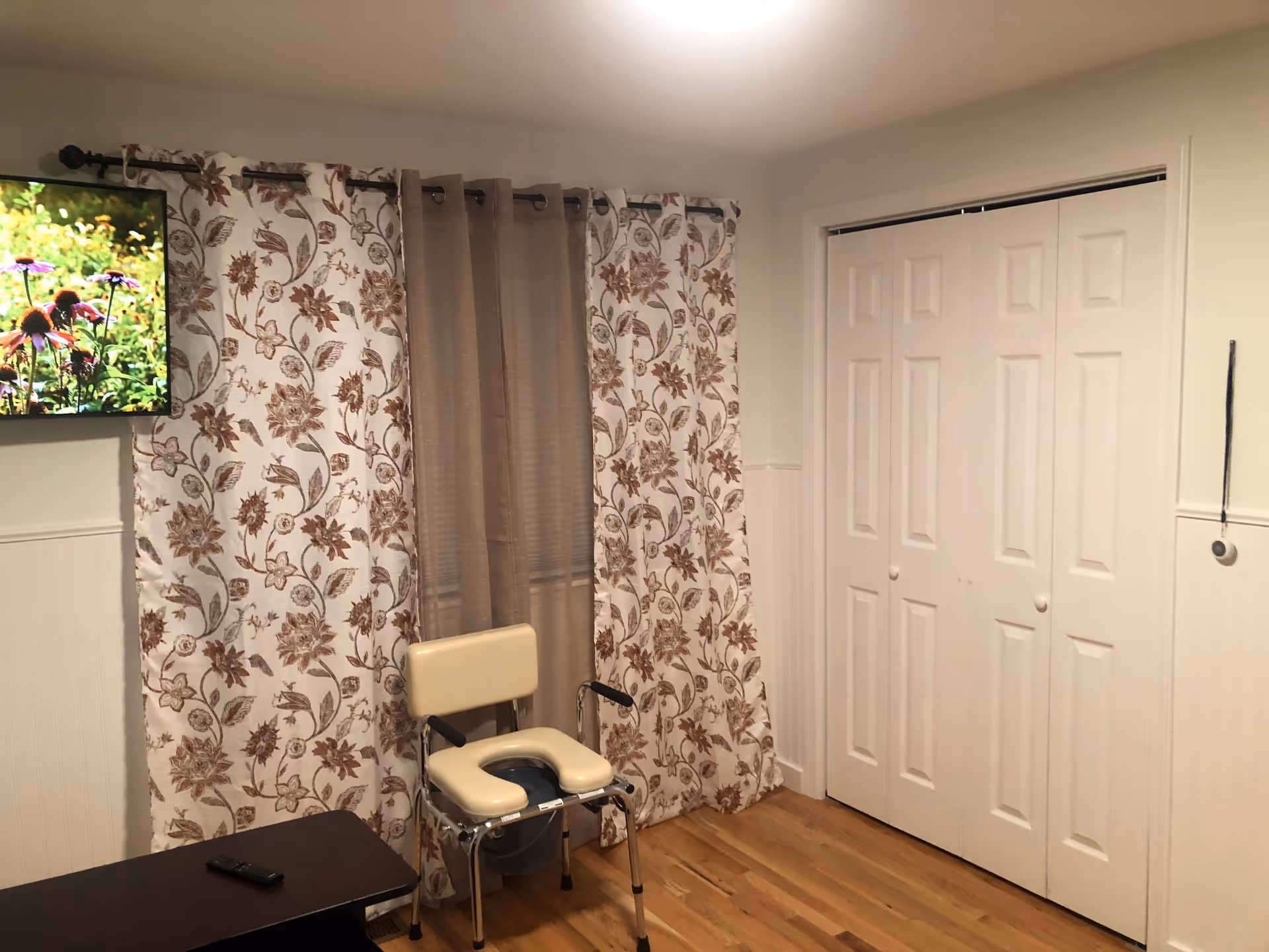 Interior room with hardwood floor, floral and beige curtains covering a window, a beige commode chair, a wall-mounted TV showing flowers, a dark wooden table with a remote control, and white double closet doors.
