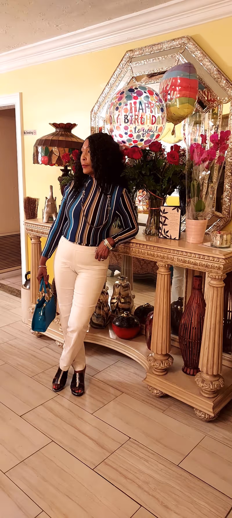 A woman with curly hair wearing a striped long-sleeve shirt and white pants is standing indoors next to an ornate wooden console table. The table holds a bouquet of red roses, a potted plant, two colorful 'Happy Birthday' balloons, and decorative items. A large decorative mirror hangs on the yellow wall behind the table.
