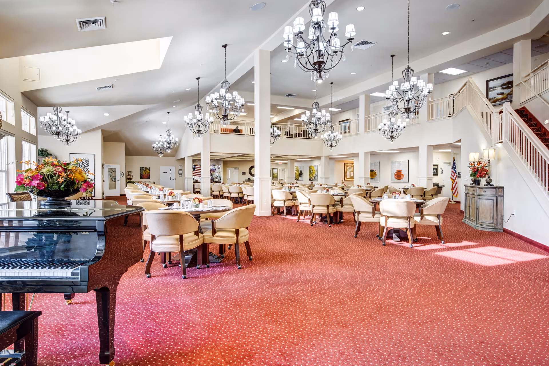 Spacious dining room with multiple round tables and beige upholstered chairs arranged neatly on a red carpet. Elegant chandeliers hang from the ceiling, and a black grand piano with a floral arrangement sits in the foreground. The room has high ceilings, large windows, and a staircase leading to an upper level.