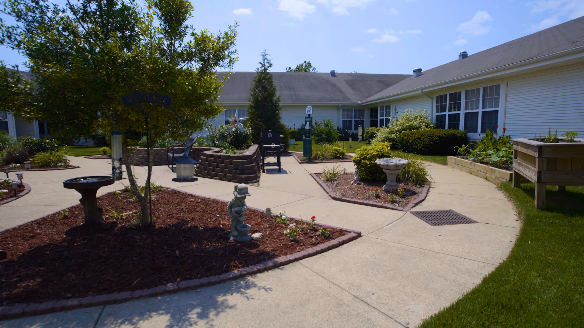 Outdoor courtyard area of a senior living facility with paved walkways, landscaped garden beds, small trees, shrubs, a birdbath, and garden decorations. The building surrounds the courtyard with large windows and a sloped roof under a clear blue sky.