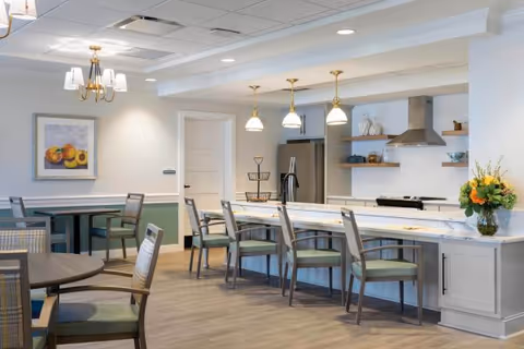 A modern dining area and kitchen with a long marble countertop featuring several chairs. The kitchen has stainless steel appliances, including a refrigerator and a range hood. Pendant lights hang above the counter, and there is a round dining table with chairs in the foreground. A framed fruit painting is on the wall, and a vase with flowers is placed on the counter.