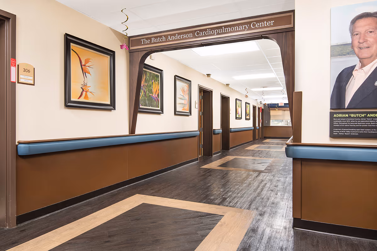 A clean, well-lit hallway in a senior living facility with brown and beige walls, blue handrails, and framed artwork on the walls. A wooden archway sign reads 'The Butch Anderson Cardiopulmonary Center.' There are several doors along the hallway and a large portrait of a man on the right side.