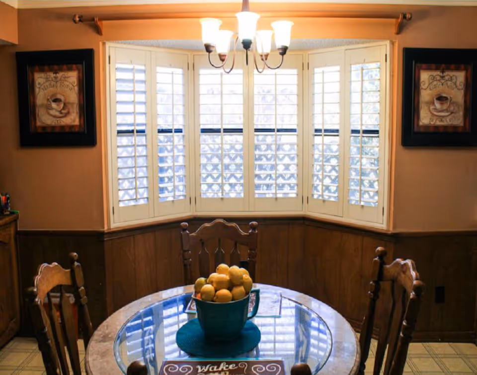 Dining area with a round glass-top table and wooden chairs under a chandelier in front of a bay window with white shutters.