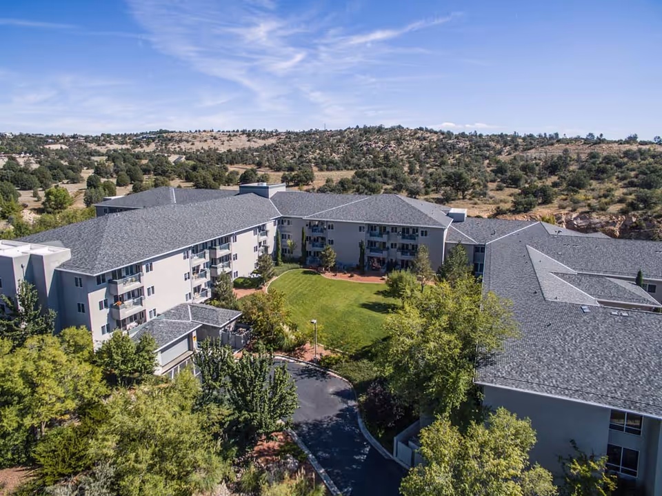 Aerial view of Las Fuentes Resort Village, showing a large multi-story residential building arranged in a U-shape around a green courtyard with trees and a lawn. The building has gray roofs and balconies, surrounded by natural landscape with hills and scattered trees under a blue sky with some clouds.