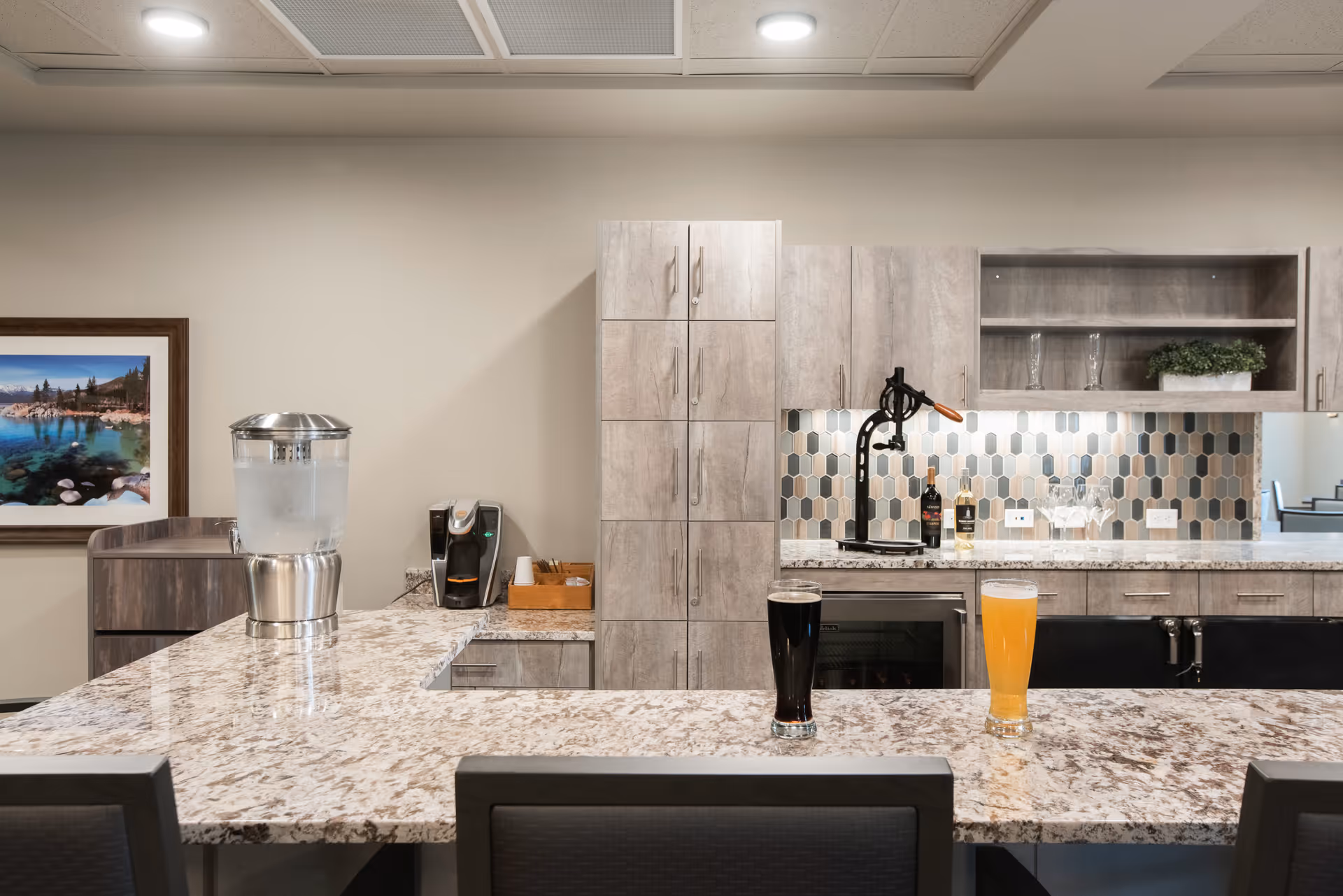 Modern kitchenette/bar area with a marble island holding two glasses of beverages, a water dispenser, coffee machine, and grey cabinetry.