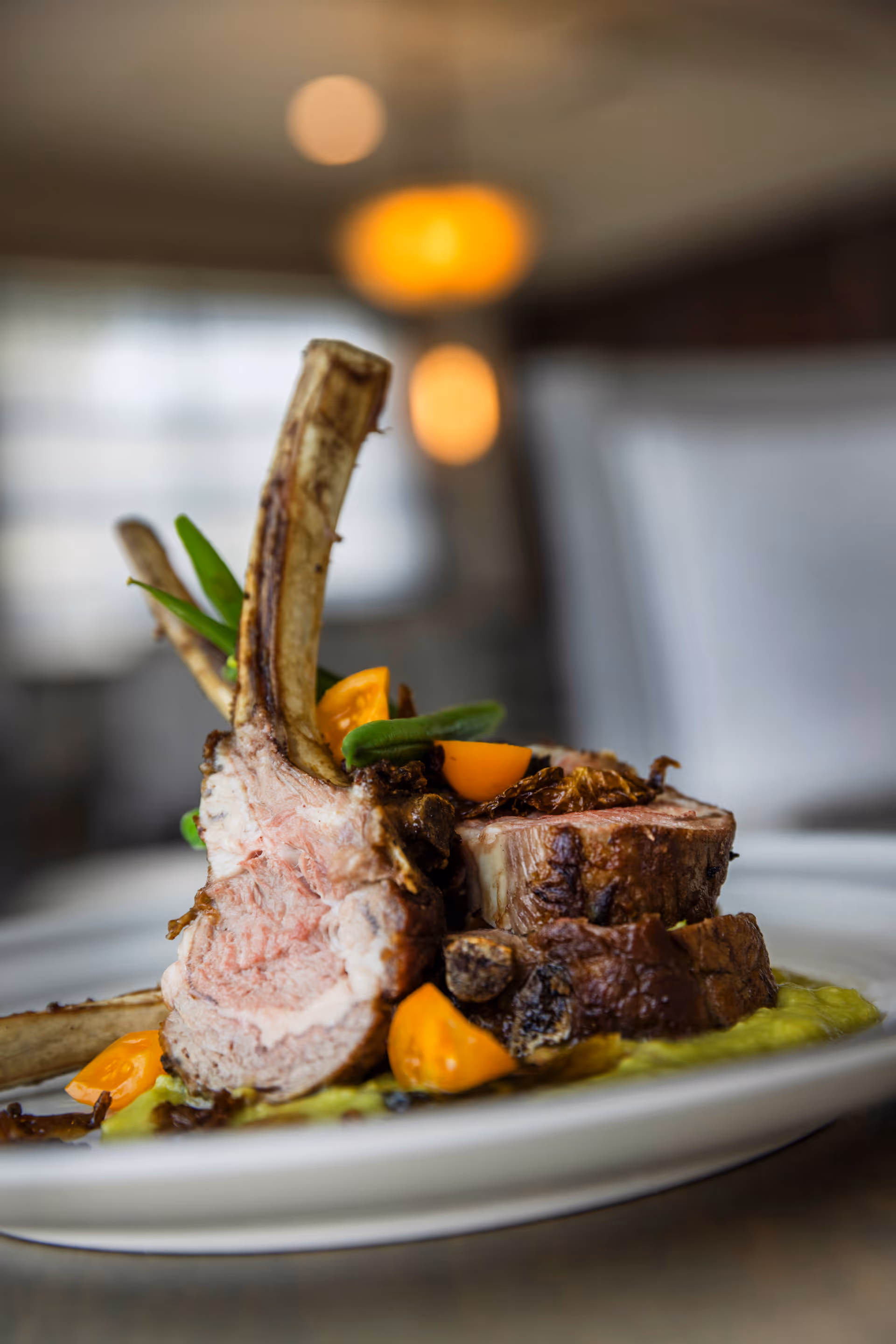 Close-up of a plated gourmet meal featuring cooked lamb chops garnished with green beans and small orange tomatoes, served on a white plate with a blurred background of a dining area.