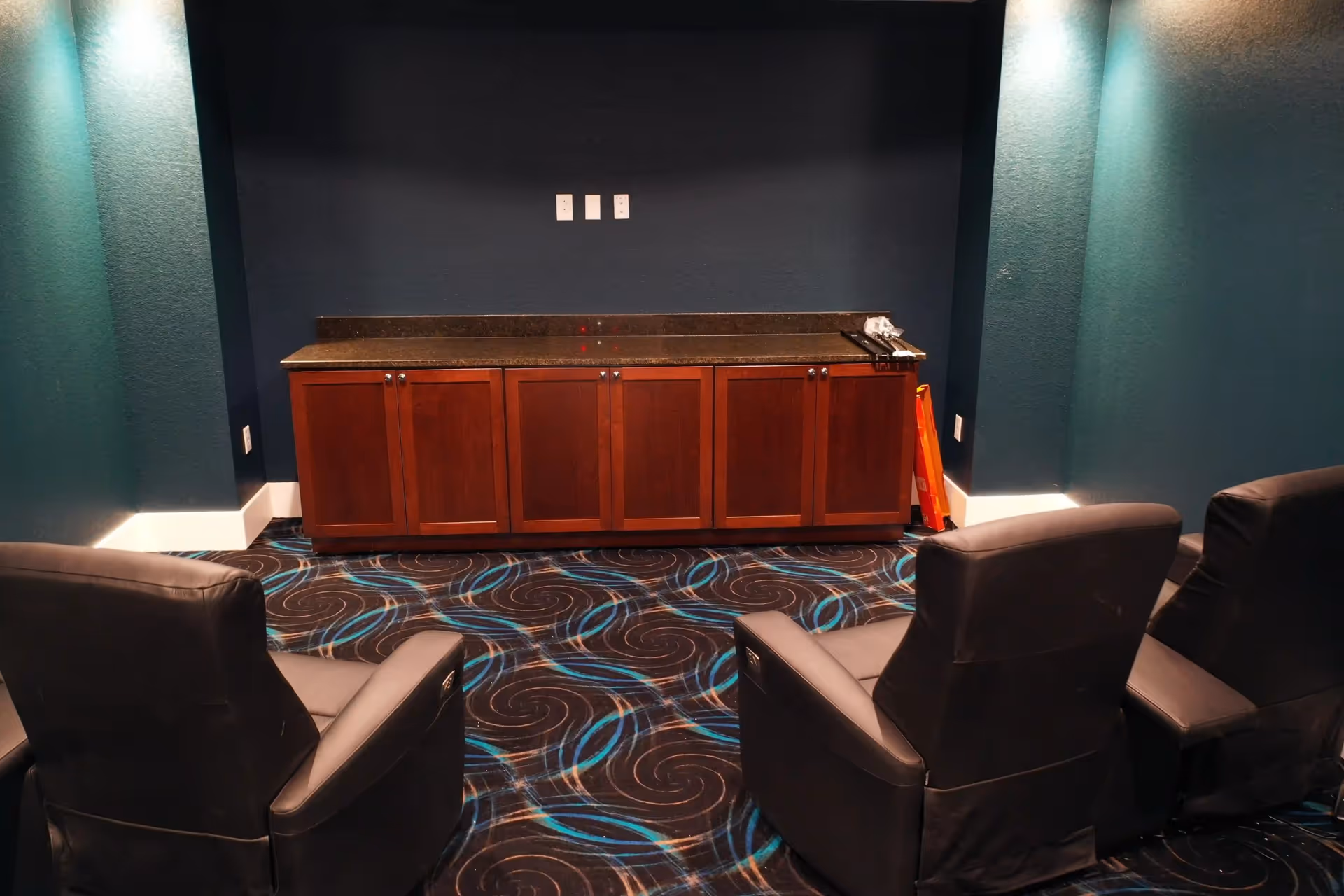 A small room with dark blue walls and patterned carpet featuring blue and brown swirls. There are three black leather recliner chairs facing a wooden cabinet with a dark countertop against the back wall. The room is softly lit with wall sconces on either side.