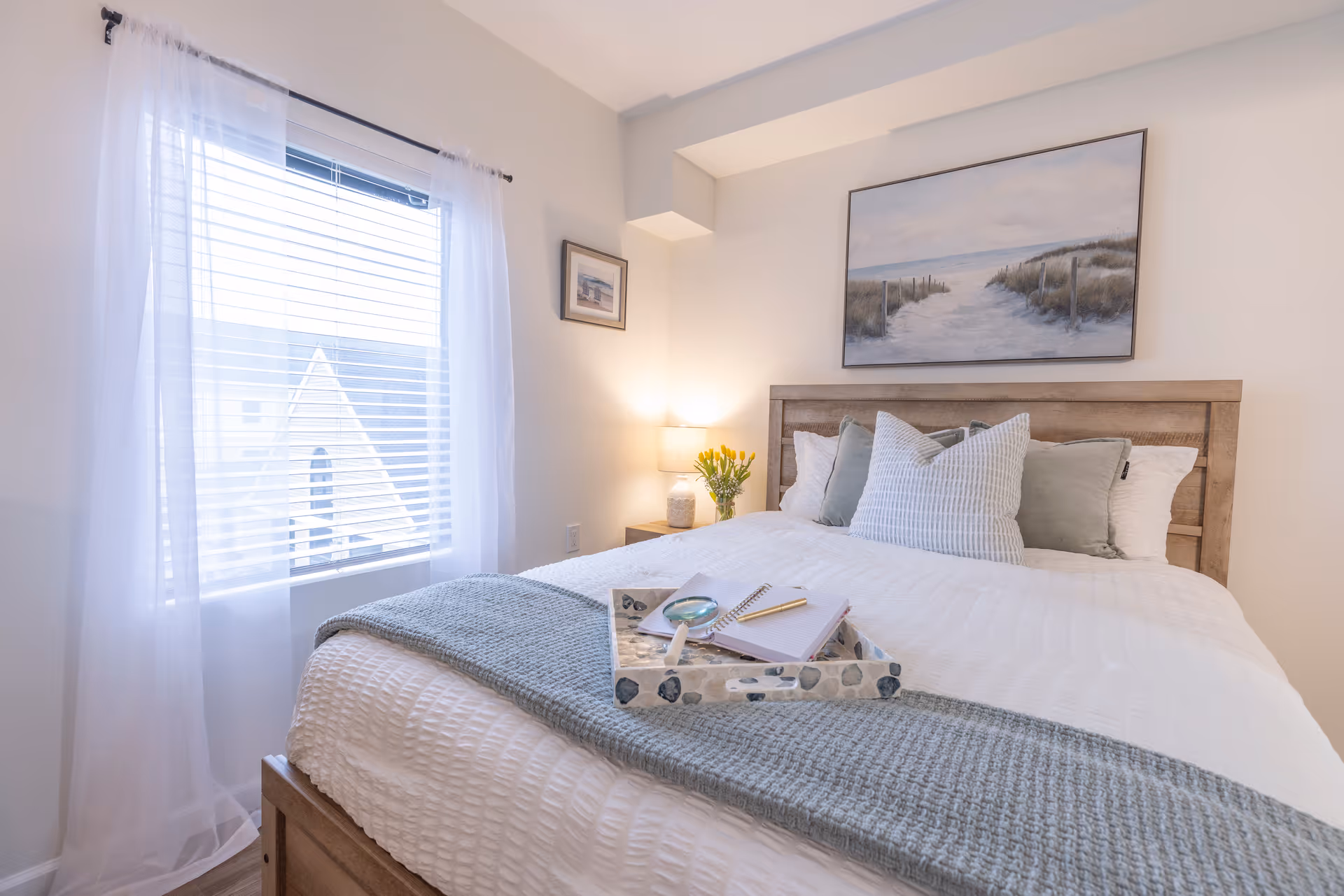 Bright, neatly made bedroom with a wooden bed, white bedding, a blue throw, and coastal artwork above the headboard.