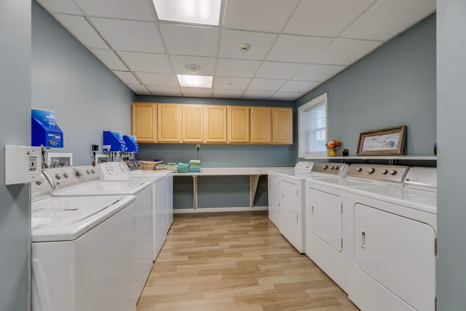 Laundry room with multiple white washing machines and dryers along both walls, light wood cabinets mounted on the far wall, a folding counter beneath the cabinets, and a window letting in natural light. The walls are painted light blue and the floor has a wood-like finish.