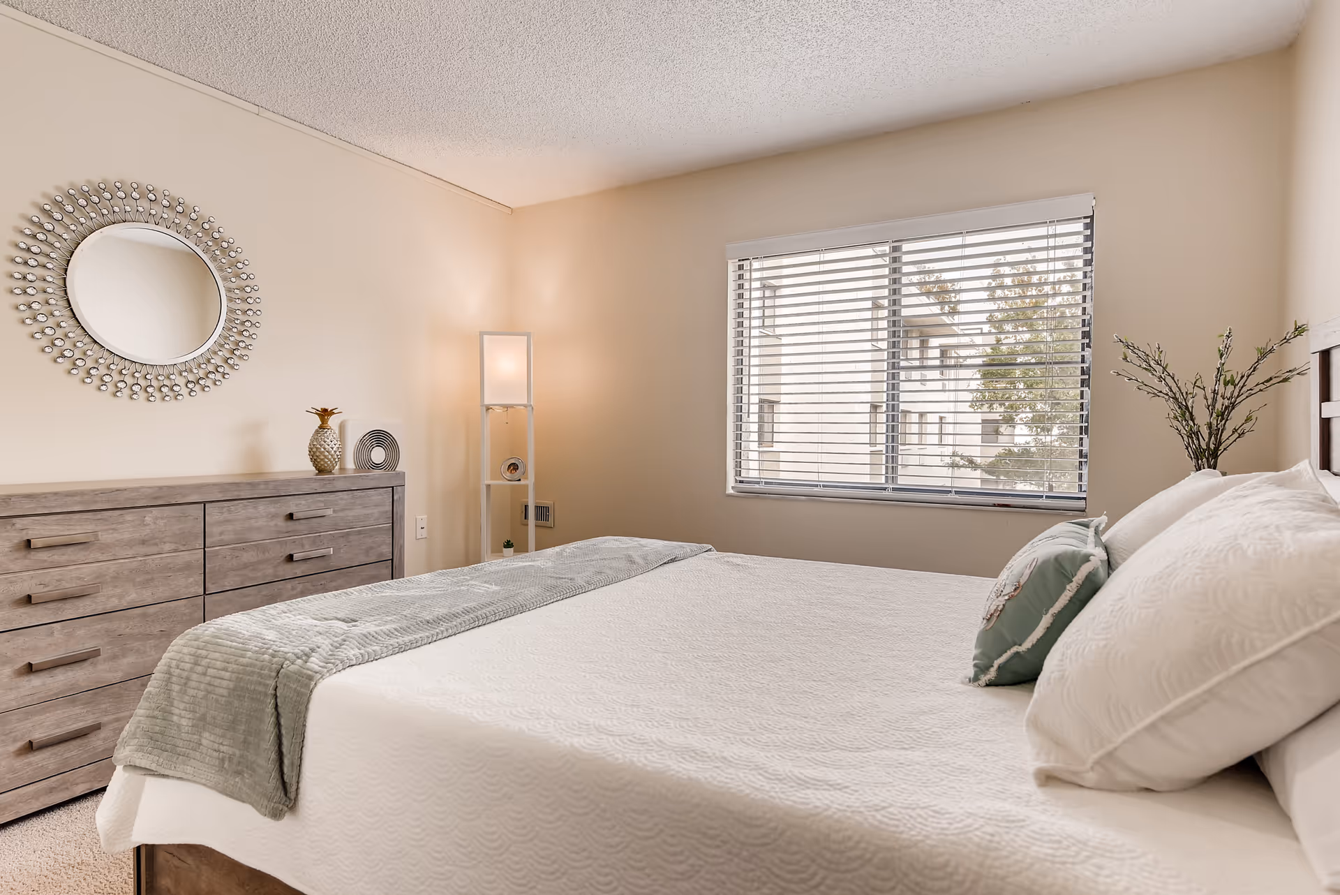 A bright and neatly arranged bedroom with a large bed covered in white bedding and a light green throw blanket. There are two pillows on the bed, one white and one green with a decorative pattern. A wooden dresser with six drawers is against the left wall, topped with decorative items including a pineapple and a circular sculpture. Above the dresser is a round mirror with a decorative frame. A tall floor lamp with three shelves stands in the corner near a window with white blinds, through which an exterior building and trees are visible. A small plant is placed on the right side of the bed near the window.