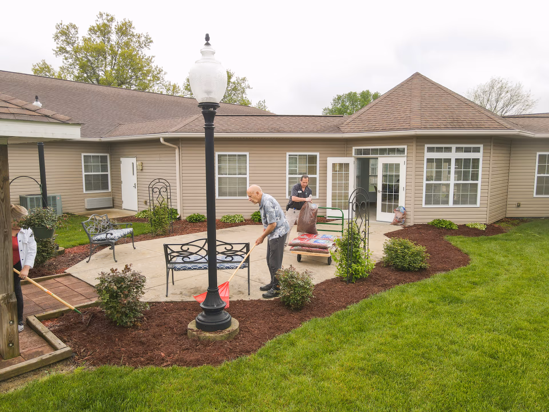 Two elderly individuals and a caregiver are tending to a garden area outside a senior living facility. One person is raking mulch around plants, while another is using a cart to transport bags of mulch. The setting includes a paved patio with benches, a lamppost, and a beige building with multiple windows and doors in the background.