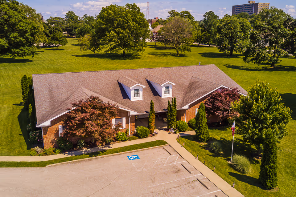 Aerial view of a single-story brick building with a gray shingled roof, surrounded by green lawns and trees. There is a small parking lot with a handicap parking space in front of the building. The building has two dormer windows on the roof and a covered entrance with shrubs and small trees around it. An American flag is visible near the entrance.