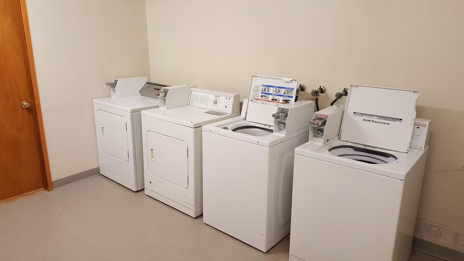 A laundry room with four white washing machines and dryers lined up against a beige wall. The machines have coin slots and some lids are open. There is a wooden door on the left side of the room.