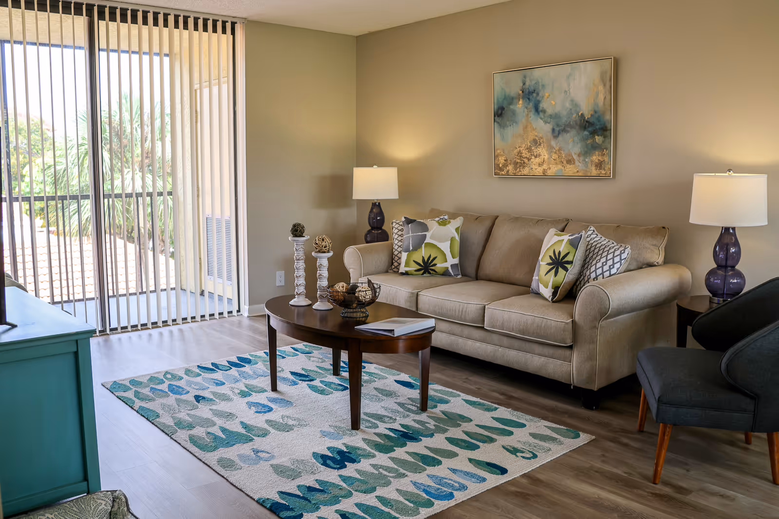 A cozy living room with a beige sofa adorned with patterned cushions, a dark wooden coffee table with decorative items, a blue and green patterned rug, two table lamps with purple bases on side tables, a dark gray armchair, and a large sliding glass door with vertical blinds letting in natural light. A colorful abstract painting hangs on the wall above the sofa.