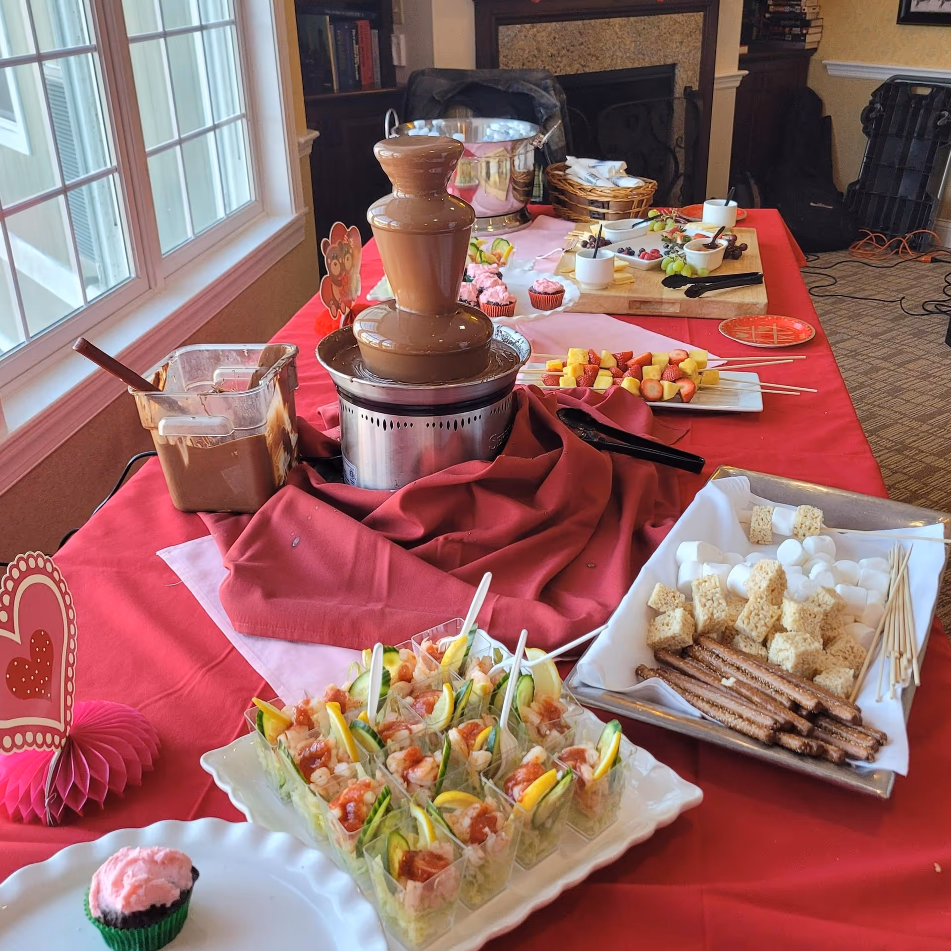 A dessert buffet table with a chocolate fountain surrounded by various treats including skewers of fruit, small cups of shrimp cocktail, marshmallows, pretzel sticks, Rice Krispies treats, and cupcakes. The table is covered with a red tablecloth and is set up near a window with natural light.