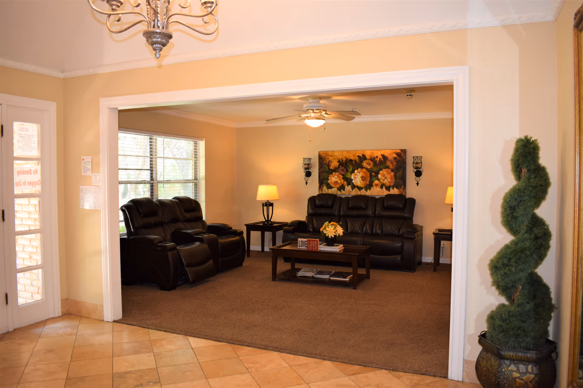 A cozy living room area with two dark leather sofas, a wooden coffee table with decorative items, two side tables with lamps, a ceiling fan with light, a large window letting in natural light, a floral painting on the wall, and a spiral-shaped topiary plant in a decorative pot near the entrance.