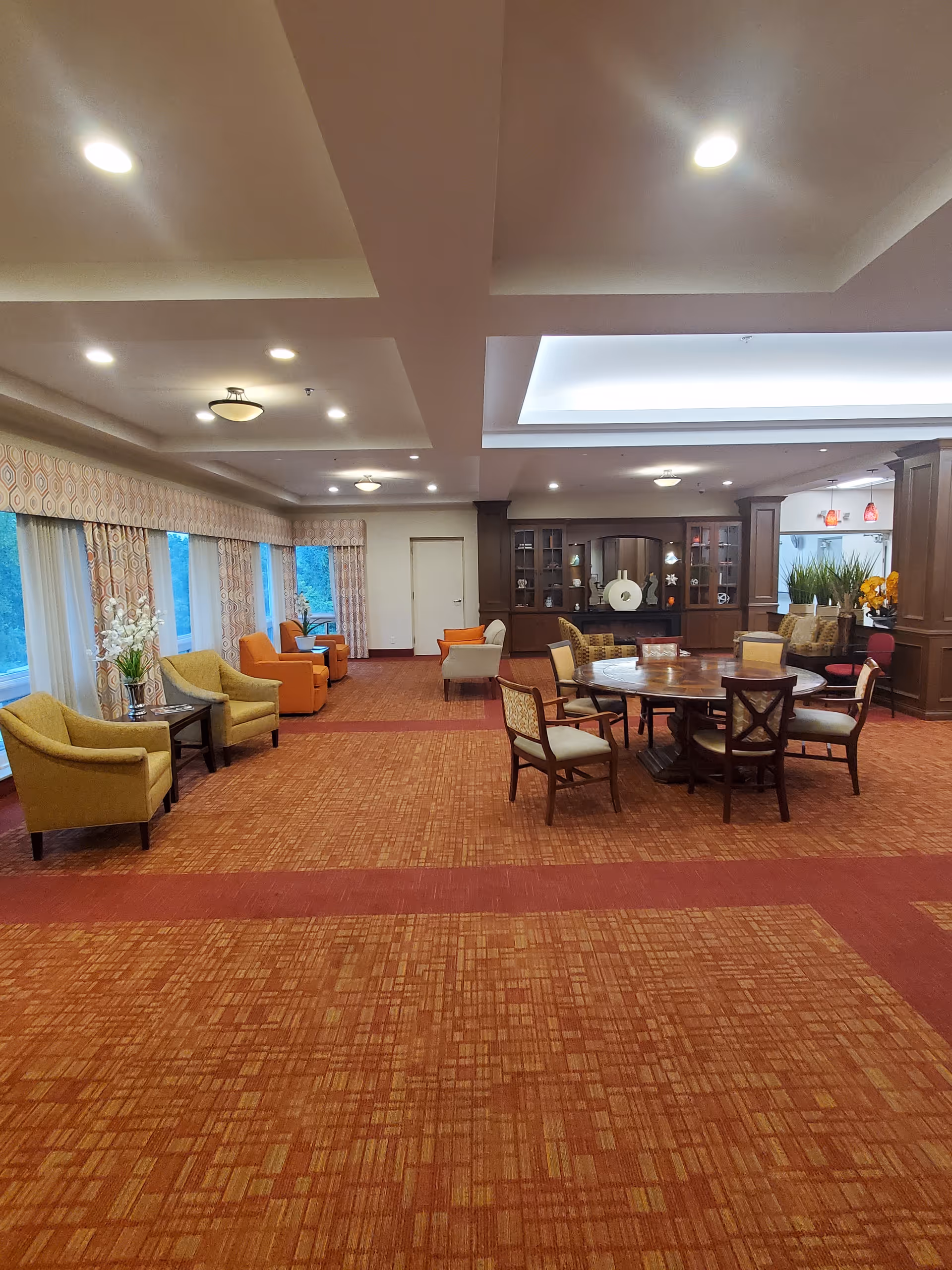 Spacious common area with patterned orange carpet, multiple seating arrangements including armchairs and a round wooden table with chairs. Large windows with patterned curtains line one side, and built-in wooden cabinets with decorative items are visible in the background. The ceiling has recessed lighting and a tray ceiling with additional lighting.