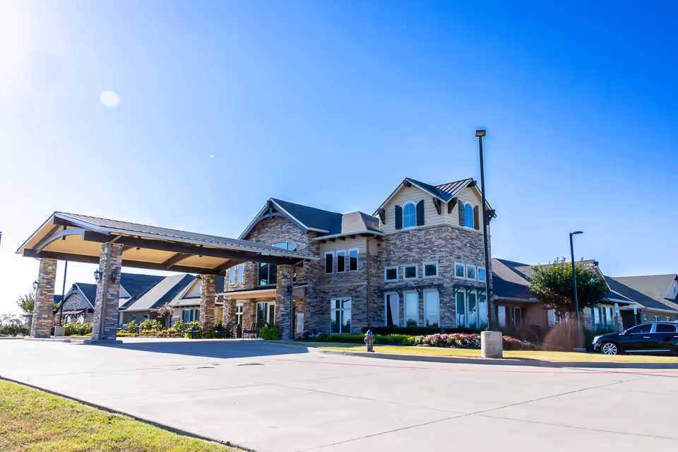 Exterior view of a large assisted living facility building with stone and brick facade, multiple windows, and a covered entrance driveway under a clear blue sky.