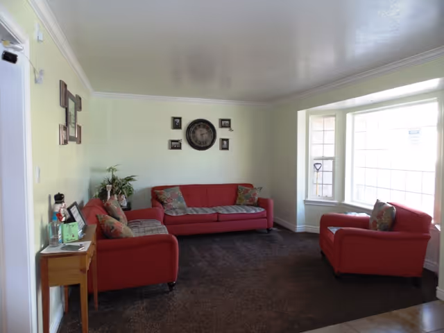 A cozy living room with light green walls, featuring a red sofa, a matching loveseat, and an armchair arranged around a dark brown carpet. There are decorative pillows on the seating, a small wooden side table with framed photos and a plant, and a large wall clock with smaller framed pictures on the wall above the sofa. A large window lets in natural light from the right side.