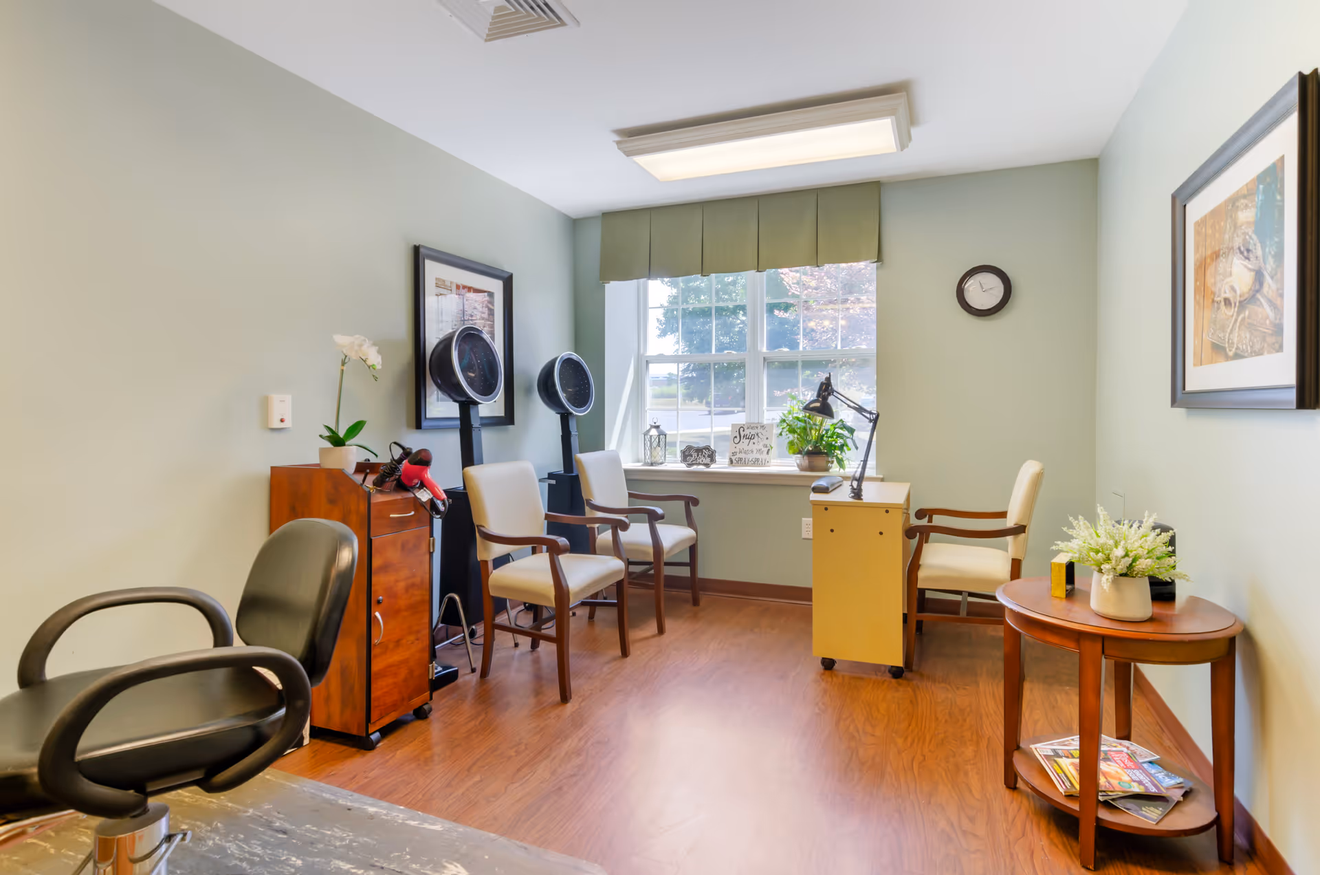 A bright and clean hair salon room with wooden flooring, light green walls, and a large window with a green valance. The room contains salon chairs, two hair dryers, a small wooden cabinet with a hairdryer on top, a small desk with a lamp and a chair, and a round wooden side table with a plant and magazines underneath. There are framed pictures on the walls and a clock above the desk.