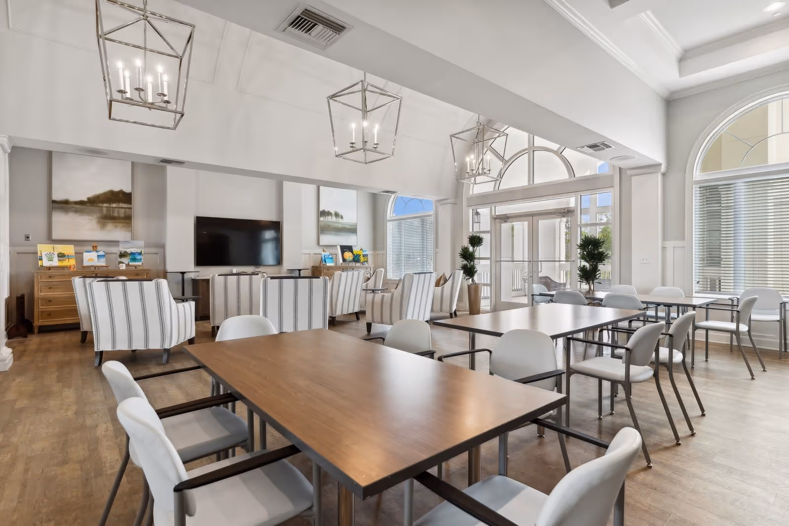 A bright and spacious common area in a senior living facility featuring multiple wooden tables with white cushioned chairs. The room has large arched windows allowing natural light to fill the space, modern hanging light fixtures, and a seating area with striped armchairs facing a wall-mounted flat-screen TV. There are decorative plants and artwork on the walls, creating a welcoming and comfortable atmosphere.