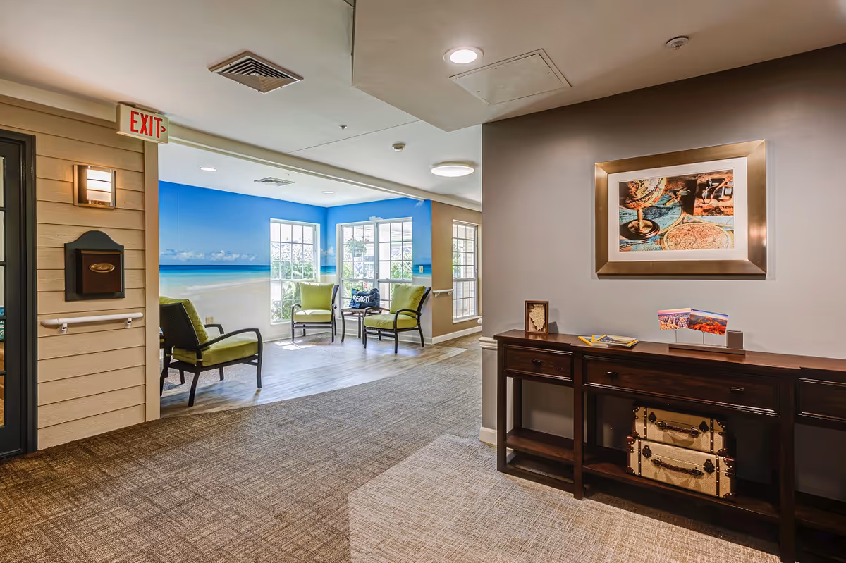 Interior view of a senior living facility hallway leading to a seating area with green cushioned chairs and a beach-themed wall mural. The hallway has carpeted flooring, a wooden console table with decorative items, and framed artwork on the wall. An exit sign is visible above a door on the left.