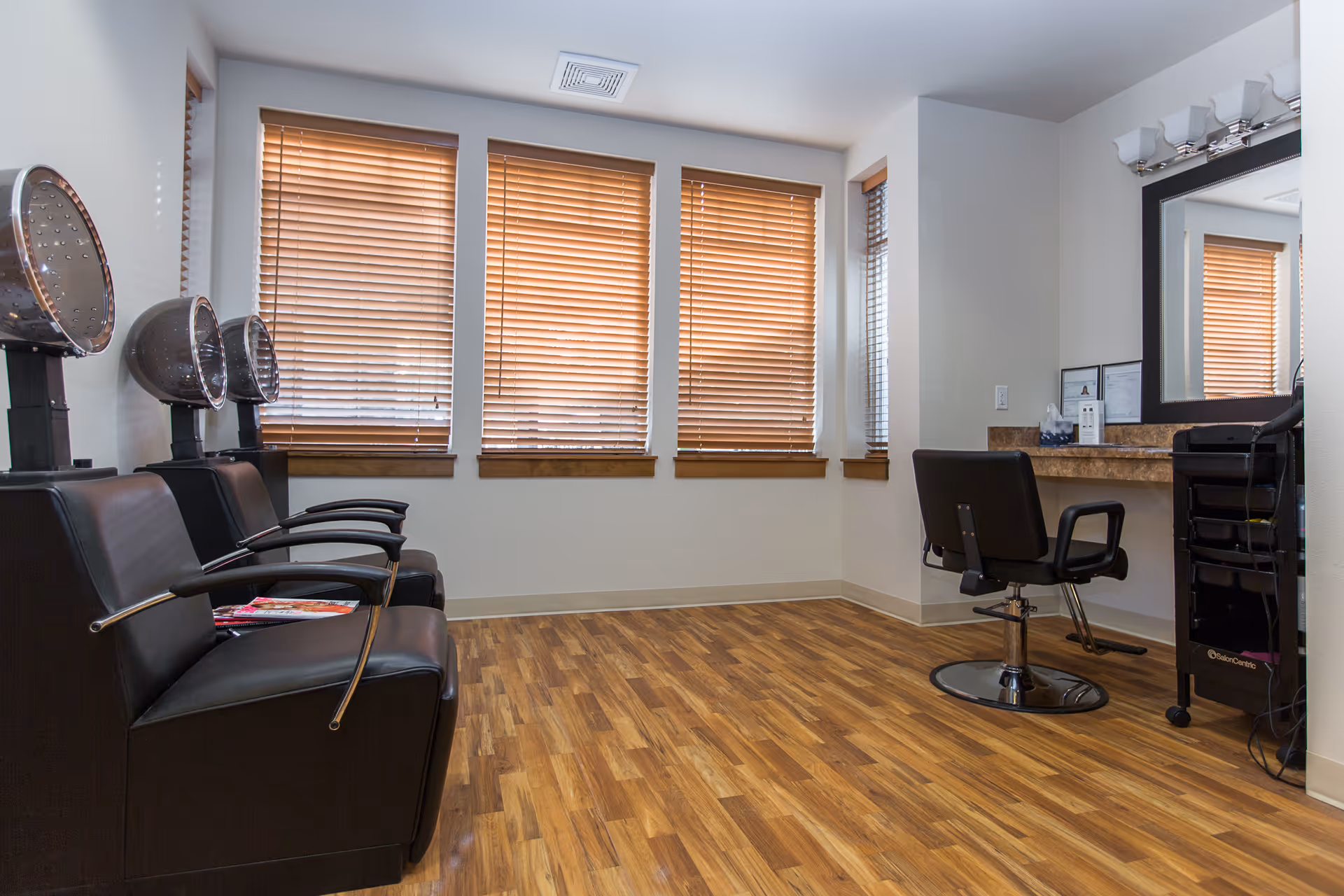 Interior view of a salon area with three black salon chairs with hair dryers on the left side and a single black styling chair in front of a large mirror on the right side. The room has wooden blinds on the windows and wood-patterned flooring.