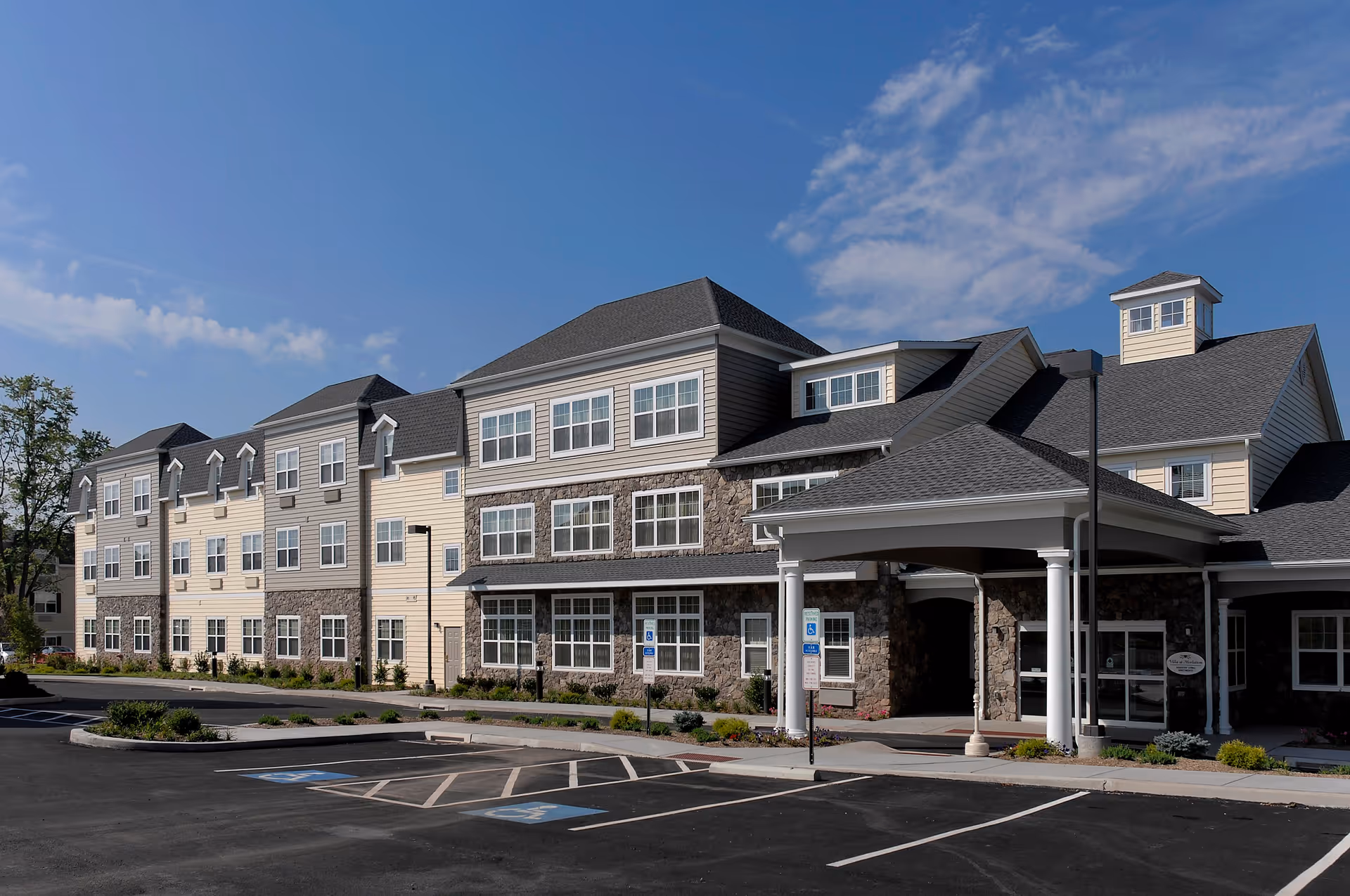 Exterior view of a multi-story senior living facility building with a covered entrance, multiple windows, stone and siding facade, and a parking lot with handicap parking spaces in front under a partly cloudy blue sky.