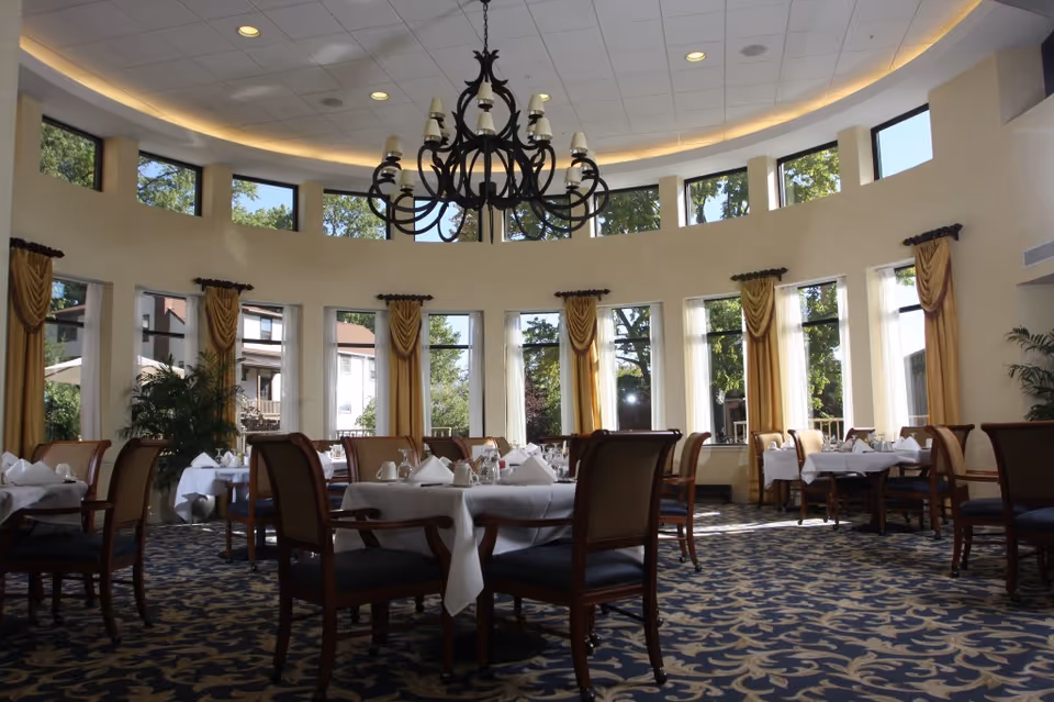 A bright and elegant dining room with large windows letting in natural light. The room features round tables covered with white tablecloths, set with napkins, glasses, and silverware. There are wooden chairs with cushioned seats around the tables. The ceiling is high and curved with a large black chandelier hanging in the center. The windows have gold curtains, and there are plants placed near the windows. Outside, trees and parts of neighboring buildings are visible.