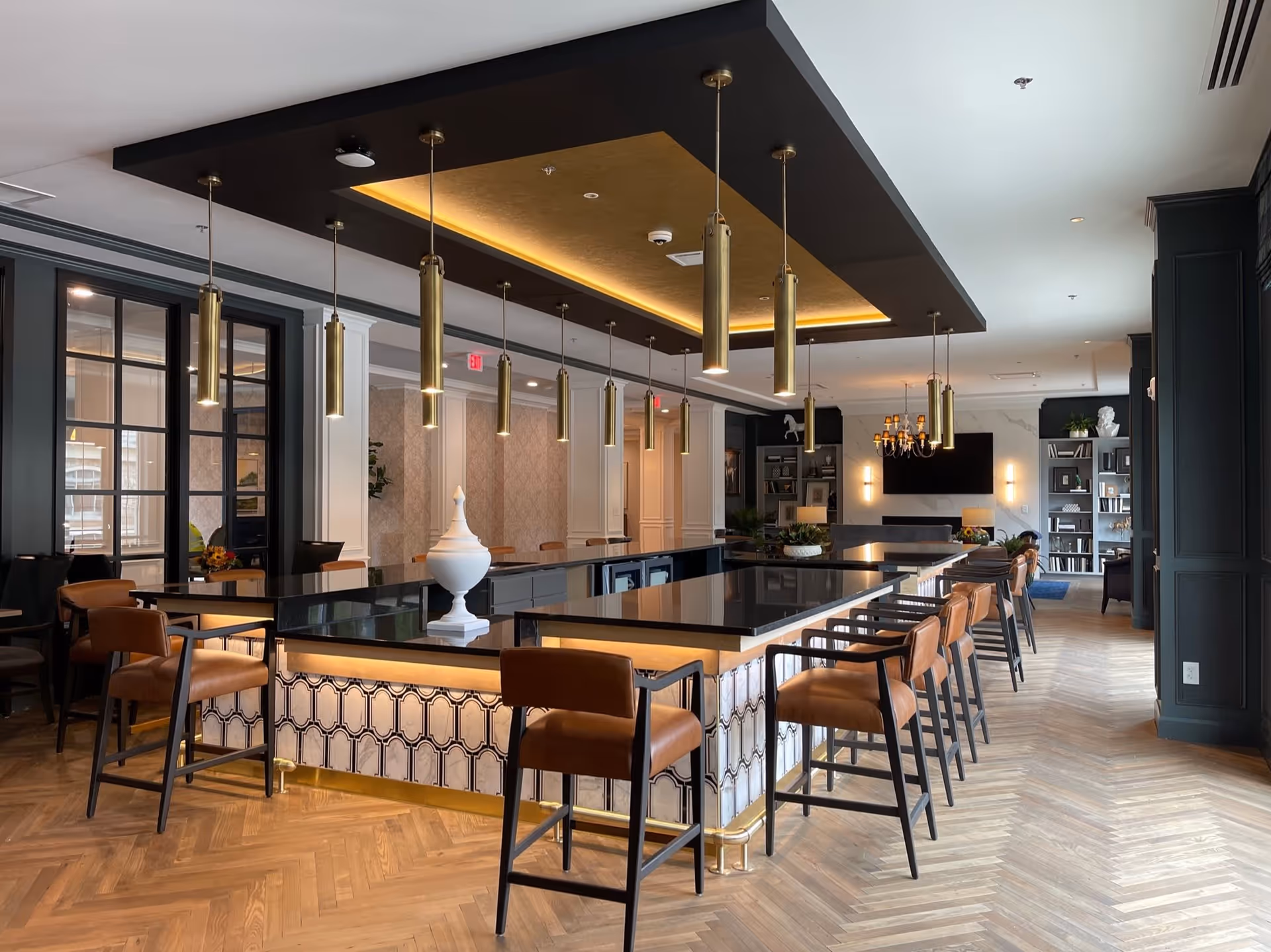 A modern and elegant common area with a large black countertop island surrounded by brown leather chairs. The ceiling above the island features a recessed lighting panel with multiple hanging gold pendant lights. The room has wood herringbone flooring, dark green paneled walls, and built-in shelves with decorative items and a TV in the background.
