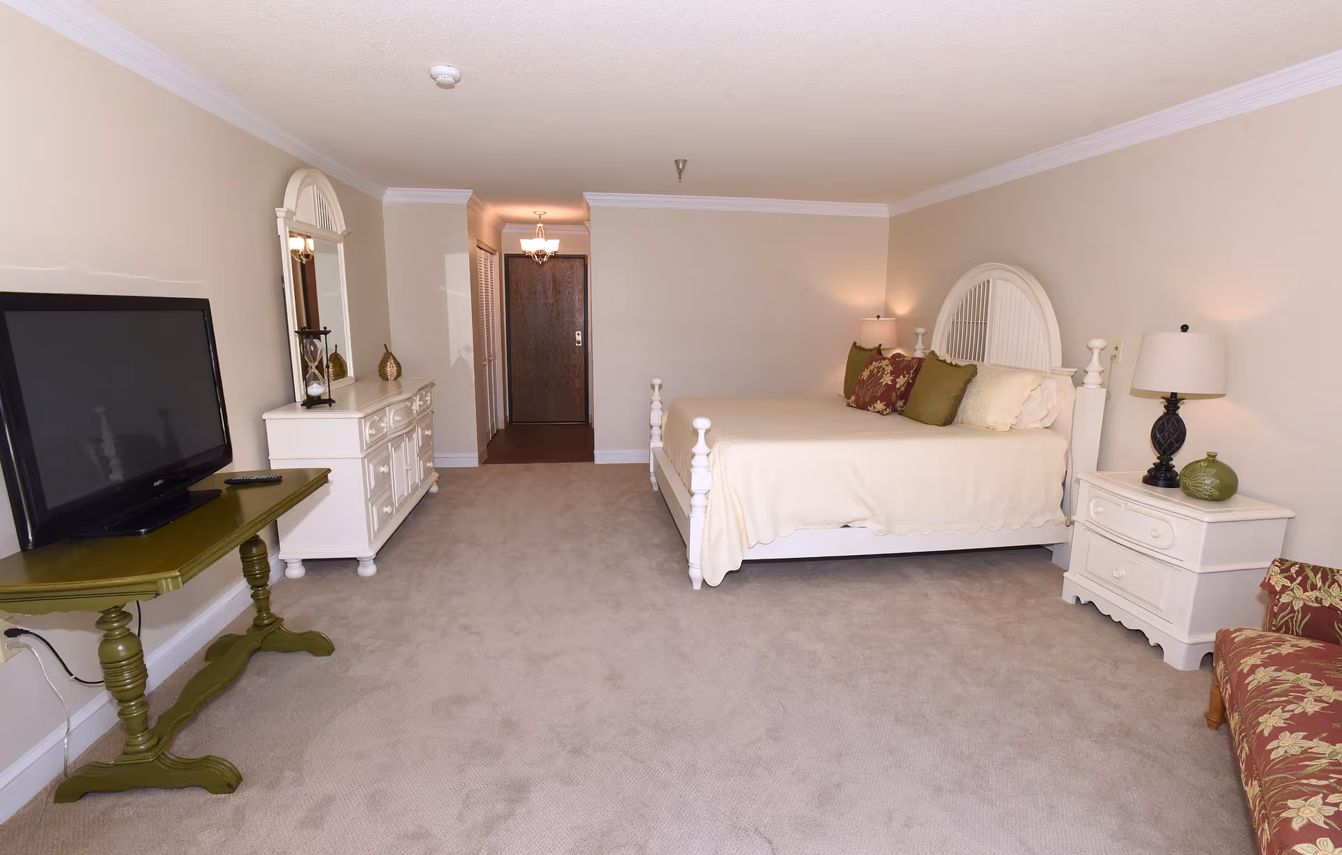 A spacious bedroom in The Residence at Eden Green featuring a white wooden bed with cream bedding and decorative pillows, a white nightstand with a lamp and decorative items, a white dresser with a mirror, a green table holding a flat-screen TV, and a floral upholstered chair. The room has beige walls and carpet, with a wooden door and hallway visible in the background.