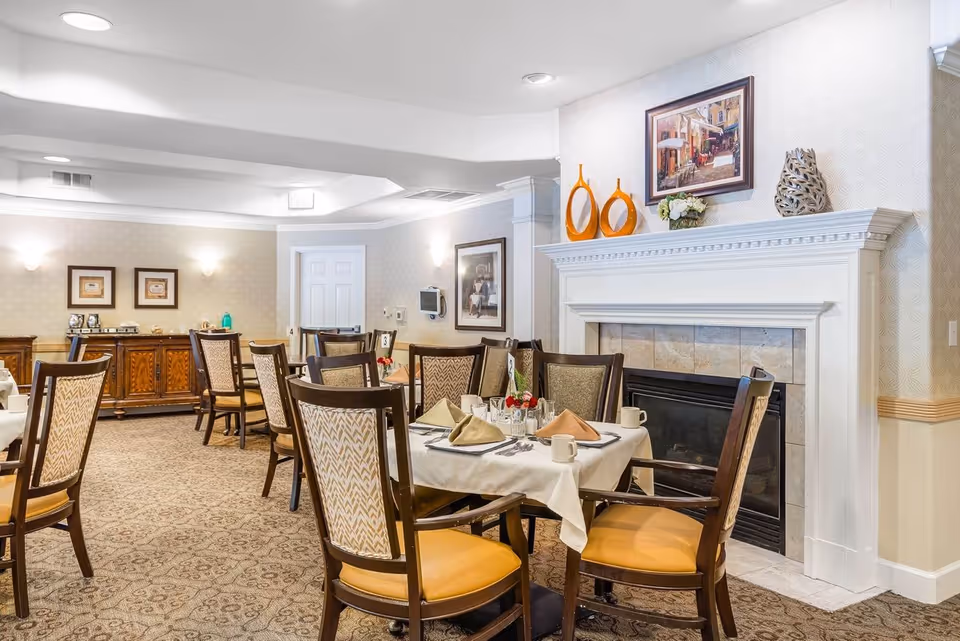 A dining room in a senior living facility with several tables set for meals. Each table has a white tablecloth, folded napkins, cups, and silverware. The room features a fireplace with decorative items on the mantel, framed artwork on the walls, and upholstered chairs with wooden frames. The carpet has a patterned design, and the lighting is soft and warm.