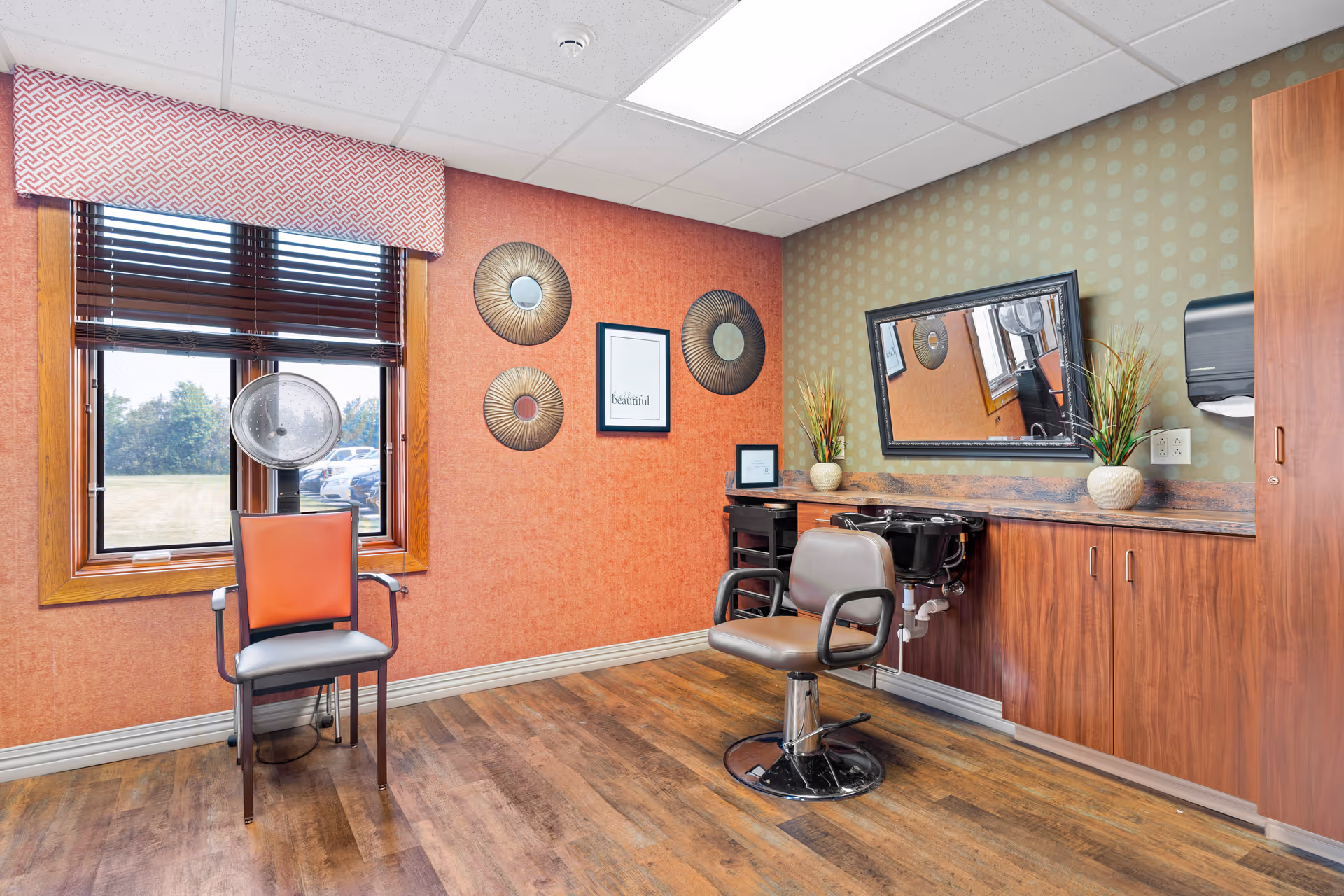 A bright and clean hair salon room with two salon chairs, a large mirror on the wall, wooden cabinets, and a window with blinds and a patterned valance. The walls are decorated with circular wall art and a framed quote. The floor is wood, and there is a hair dryer attached to one chair.