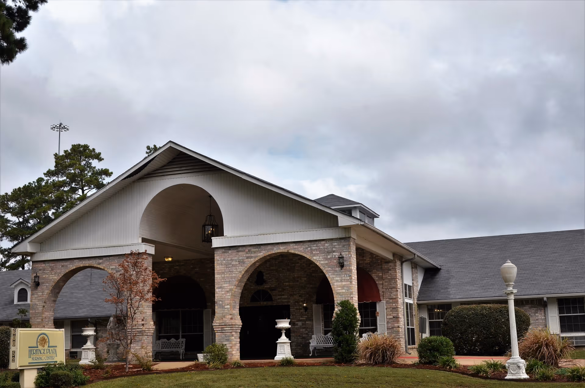 Exterior view of Heritage Plaza Nursing Center showing a brick building with arched entryways, a covered porch area with benches, landscaped bushes and trees, and a cloudy sky overhead.