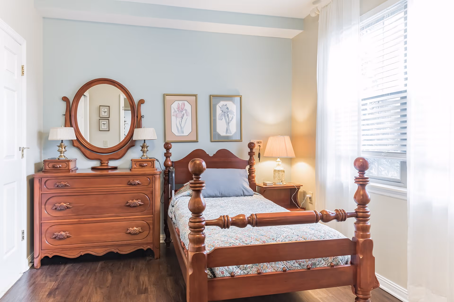 A bright bedroom with a wooden bed featuring a patterned bedspread and a gray pillow. Next to the bed is a wooden nightstand with a lamp. On the left side of the room is a wooden dresser with an oval mirror and two small lamps. Two framed floral artworks hang on the light blue wall above the bed. A window with white blinds and sheer curtains allows natural light to fill the room.