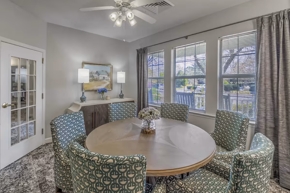 A dining room with a round wooden table surrounded by six patterned upholstered chairs. The room has three large windows with blinds and curtains, a ceiling fan with lights, a sideboard with two table lamps, a vase with flowers, and a framed painting on the wall. The floor is carpeted and there is a glass-paneled door on the left.