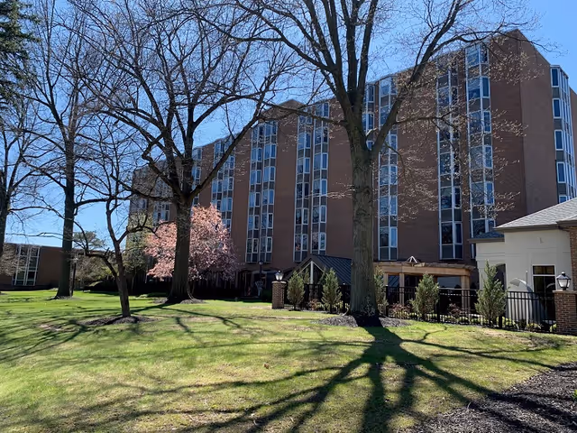 Exterior view of a multi-story brick building with large windows, surrounded by leafless trees and a green lawn under a clear blue sky. A flowering tree with pink blossoms is visible near the building.