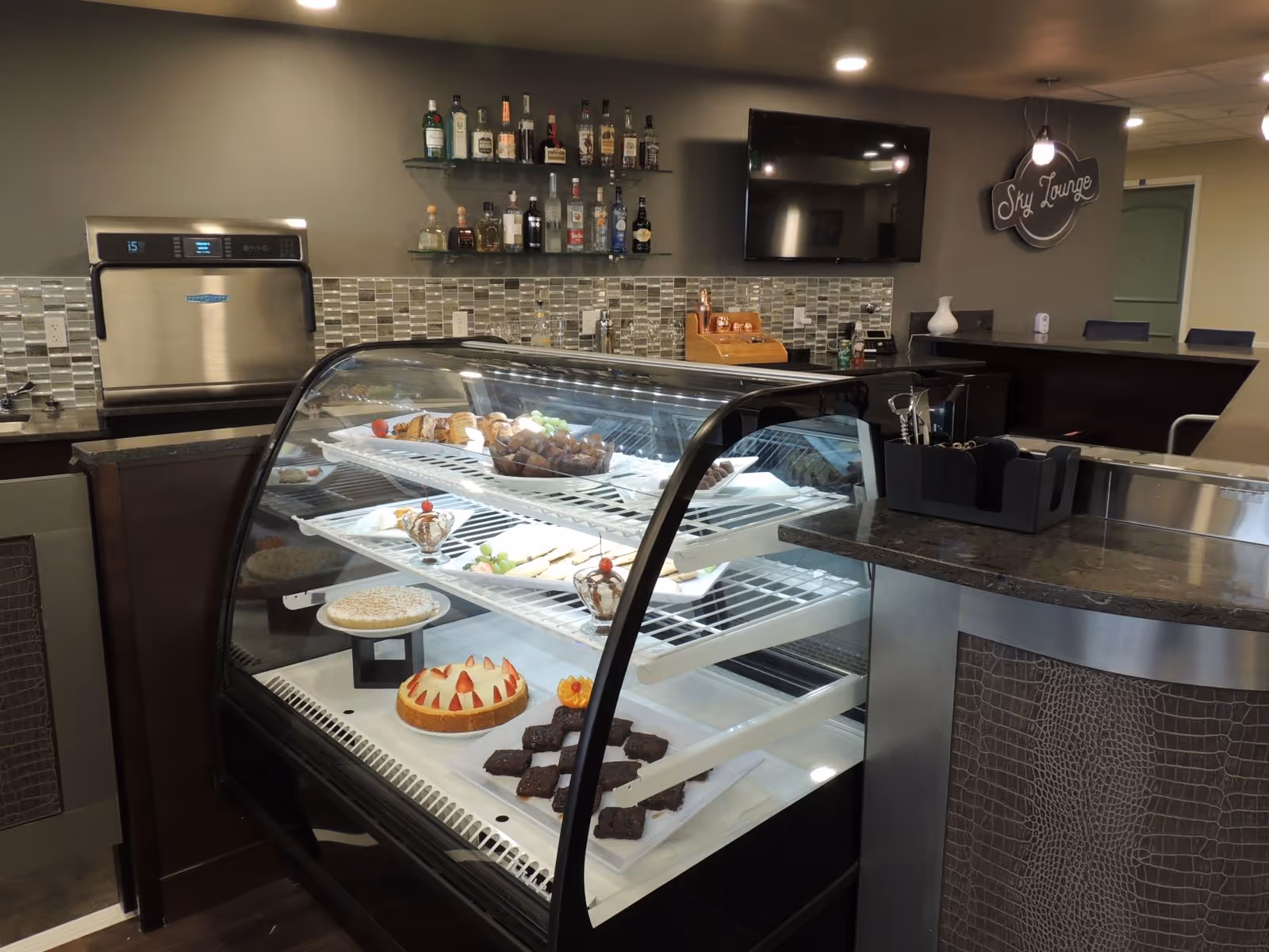Pastry display case filled with desserts in a lounge-style cafe and bar area with bottles, a TV, and a 'Sky Lounge' sign.