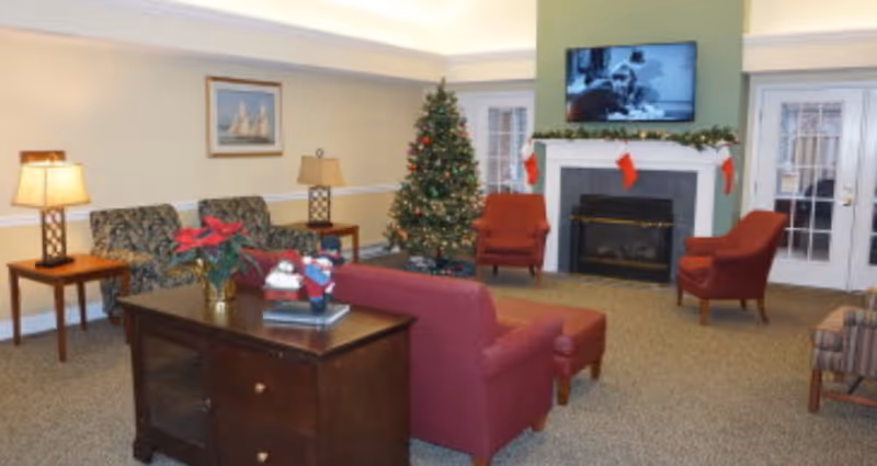 A cozy living room decorated for Christmas with a Christmas tree, stockings hanging on a fireplace mantel, a flat-screen TV above the fireplace, several armchairs and sofas, a wooden side table with a lamp, and a wooden cabinet with holiday decorations on top.