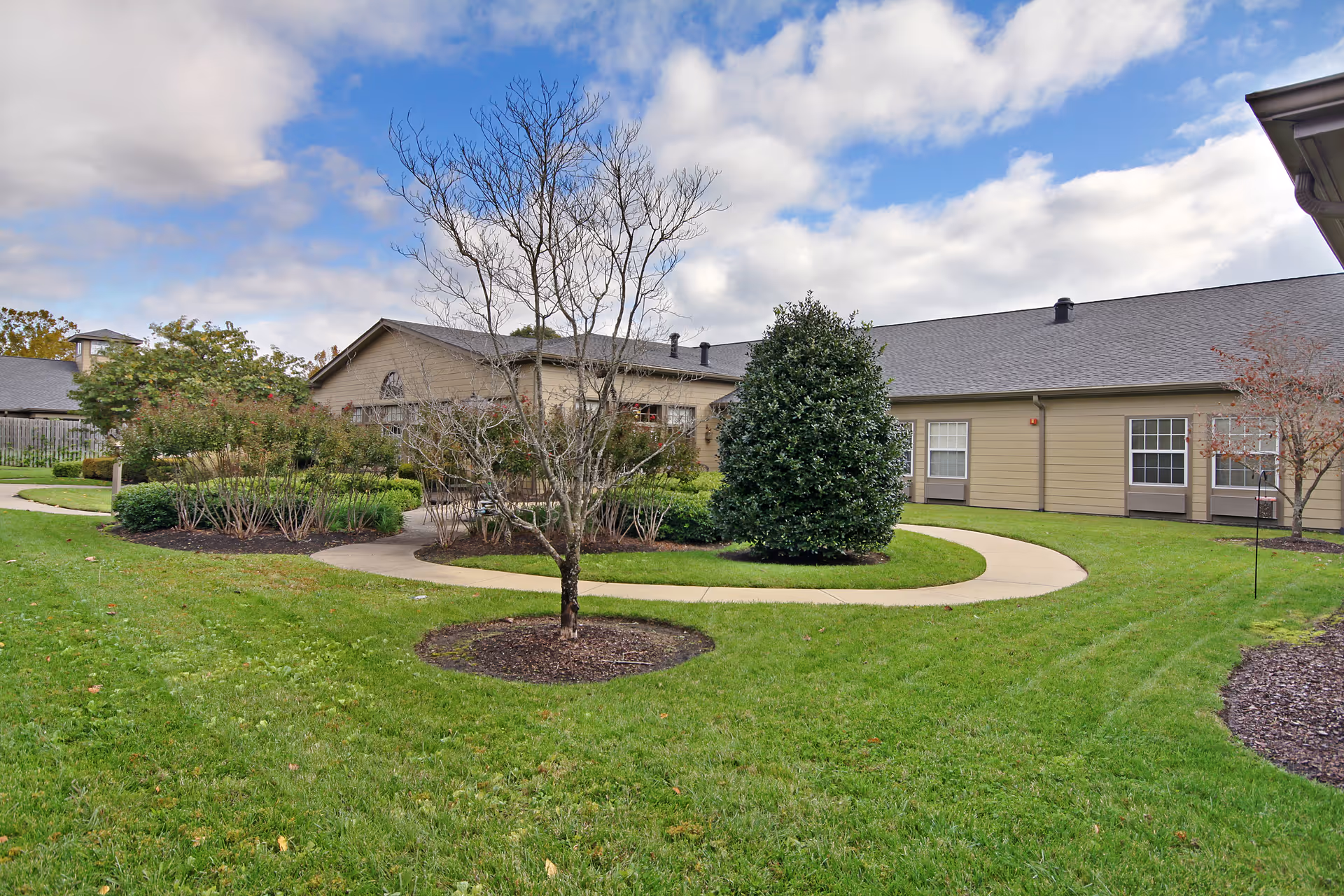 Outdoor garden area with a curved concrete pathway surrounded by green grass, bushes, and trees in front of a beige single-story building under a partly cloudy sky.