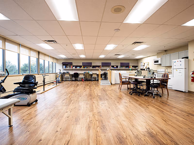 A spacious room with wooden flooring and large windows along one wall letting in natural light. The room contains exercise equipment including a recumbent bike and parallel bars on the left side. At the back, there are desks with chairs and various supplies, and on the right side, there is a round table with chairs and kitchen appliances including a refrigerator, microwave, and cabinets.