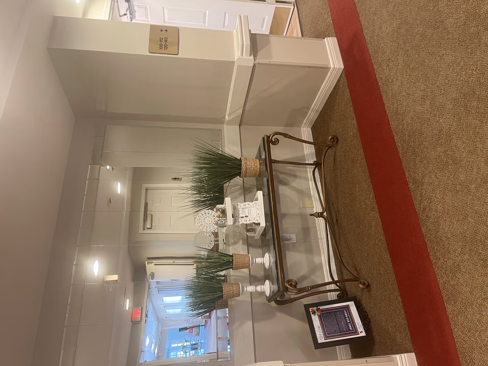 Interior lobby hallway with a glass-topped console table holding potted grasses and decor in front of a large mirror and a red carpet runner.