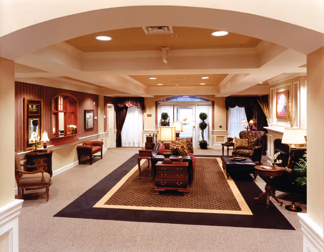 A spacious and elegantly decorated living room area with a central seating arrangement including sofas and armchairs around a coffee table on a patterned rug. The room features warm lighting from table lamps, framed artwork on the walls, and large windows with curtains. There is a fireplace on the right side and decorative plants near the entrance at the back.