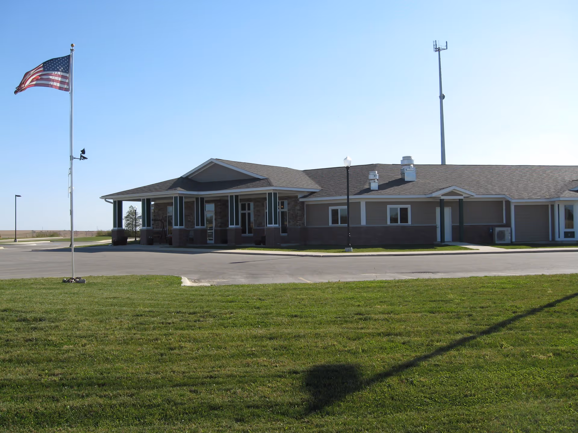 Single-story assisted living building front with an American flag on a flagpole, a paved driveway, and a grassy lawn.