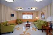 A cozy living room with green upholstered sofas and armchairs arranged around a wooden coffee table. The room has patterned wallpaper, a window with an orange valance, ceiling lights, and framed artwork on the wall. There are also two side tables with lamps.