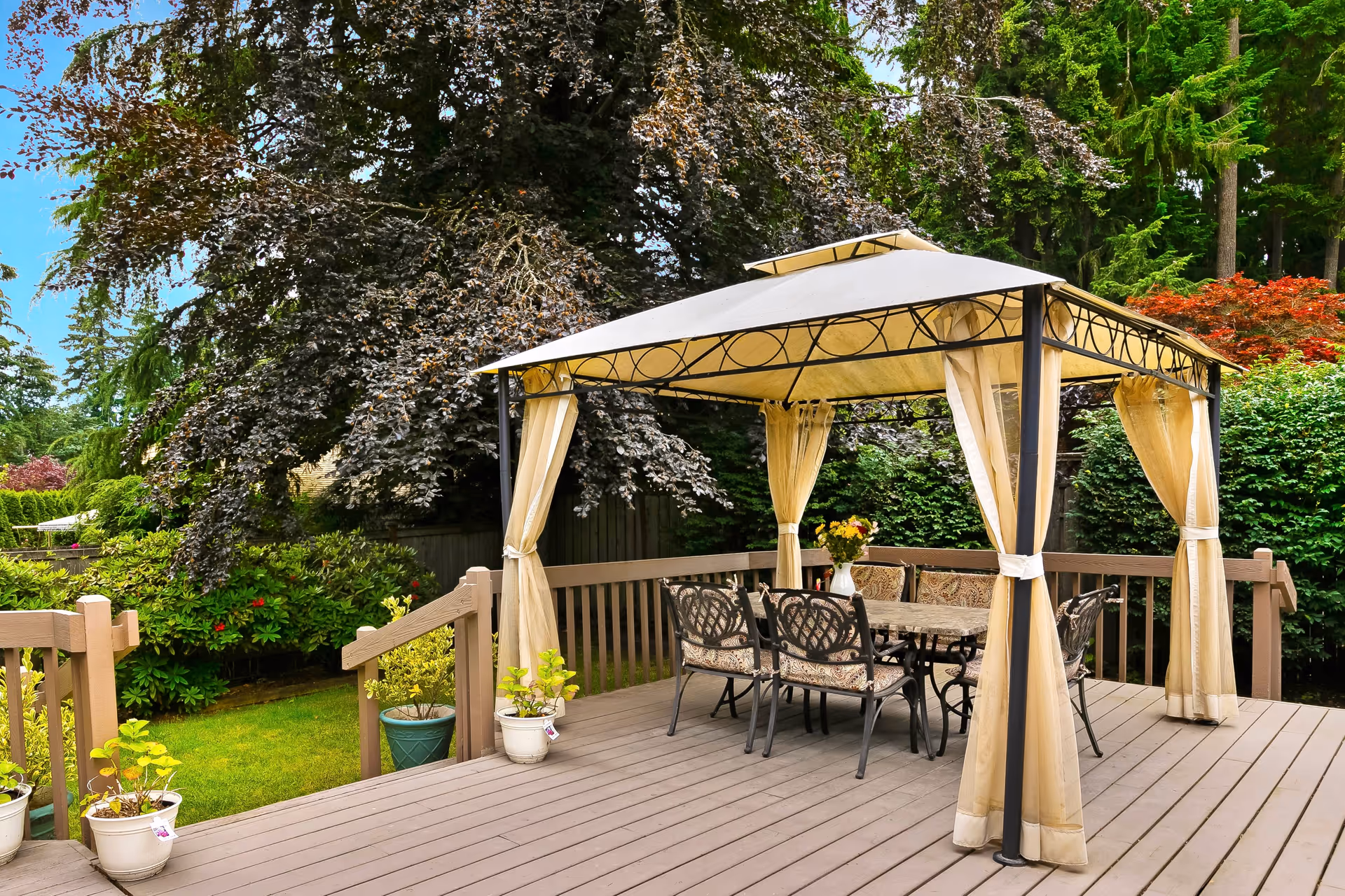Outdoor wooden deck area with a beige canopy gazebo covering a dining table and six cushioned chairs. The deck is surrounded by lush green trees and bushes, with several potted plants placed along the railing.