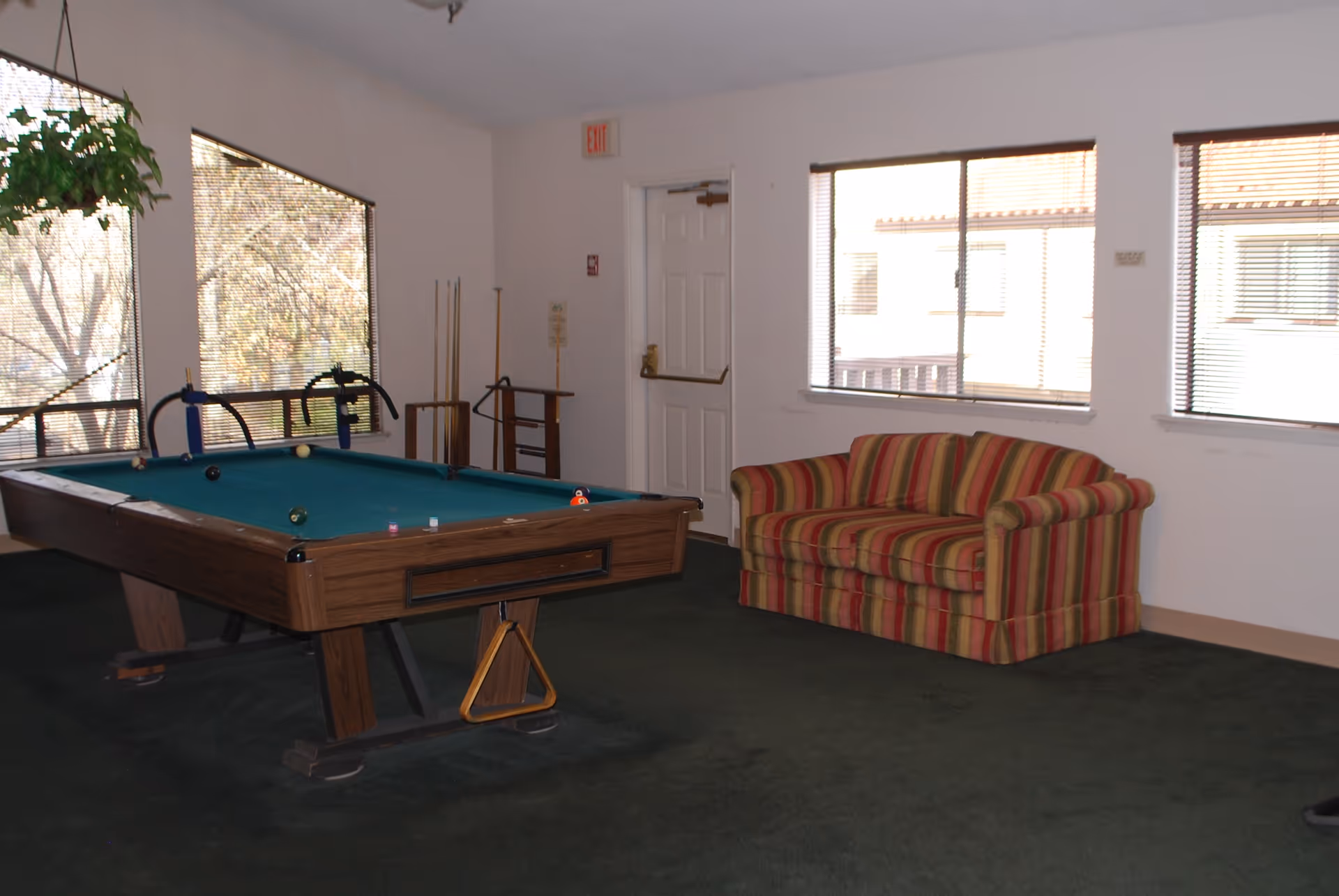 A recreational room with a pool table in the center, a striped red and yellow couch against the wall, large windows letting in natural light, and pool cues standing in a rack near the door.