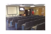 Small auditorium-style room with rows of theater seats facing a projector screen and a wall-mounted TV, with a vending machine and a person at the back.