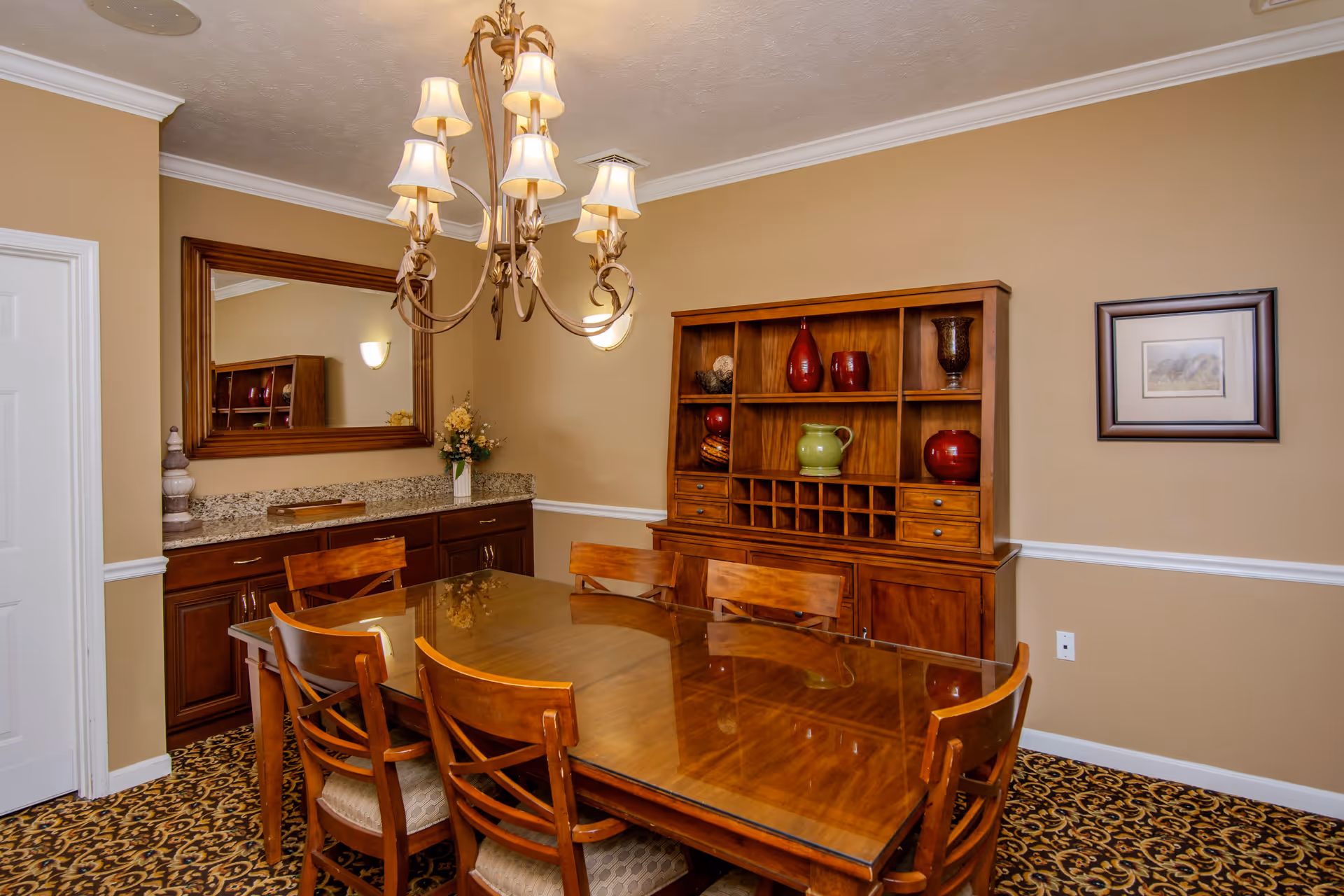 A dining room with a wooden table and six matching chairs. A wooden cabinet with decorative vases and a wine rack is against the wall. A large mirror hangs above a granite countertop with dark wooden cabinets below. A chandelier with multiple lampshades hangs above the table. The walls are painted beige with white trim, and the floor has a patterned carpet.